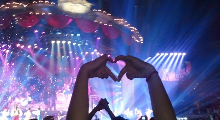 Stage at a concert seen from the audience, with an audience member holding their hands in the air in the shape of a heart